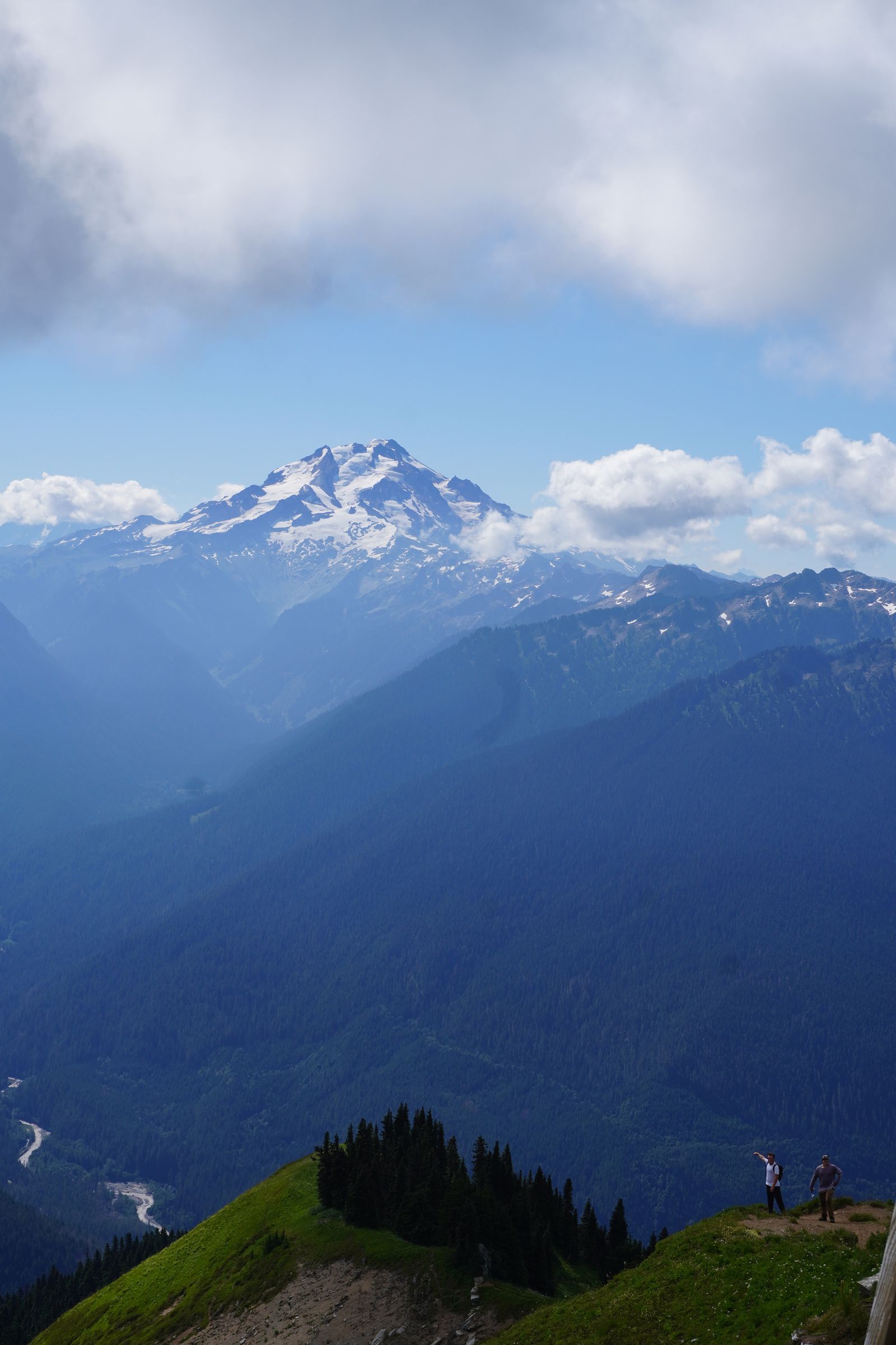 Two hikers on a grassy trail below a snow-covered peak, North Cascades