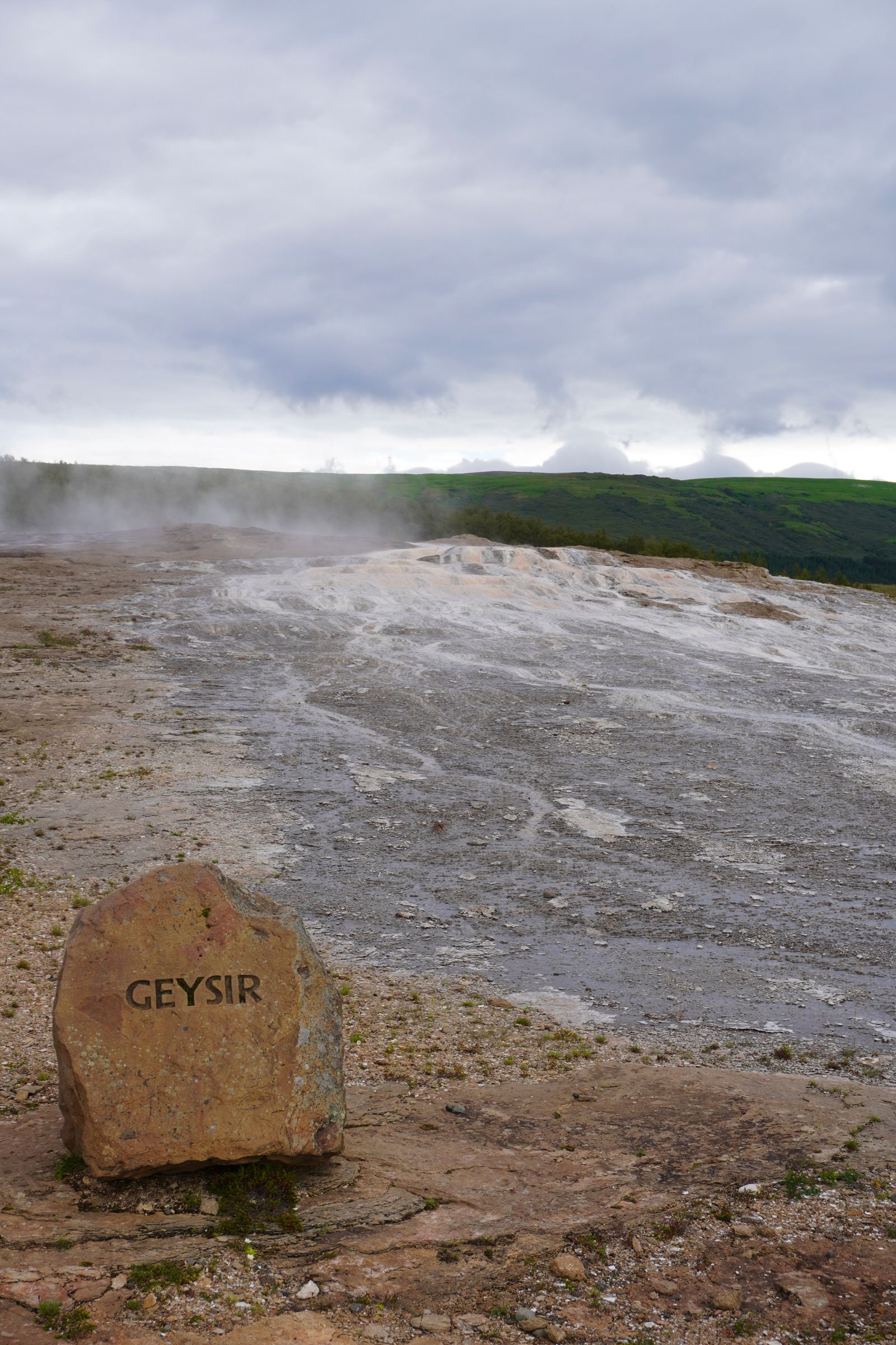 Steaming ground beyond a stone marker below green hills, Bláskógabyggð