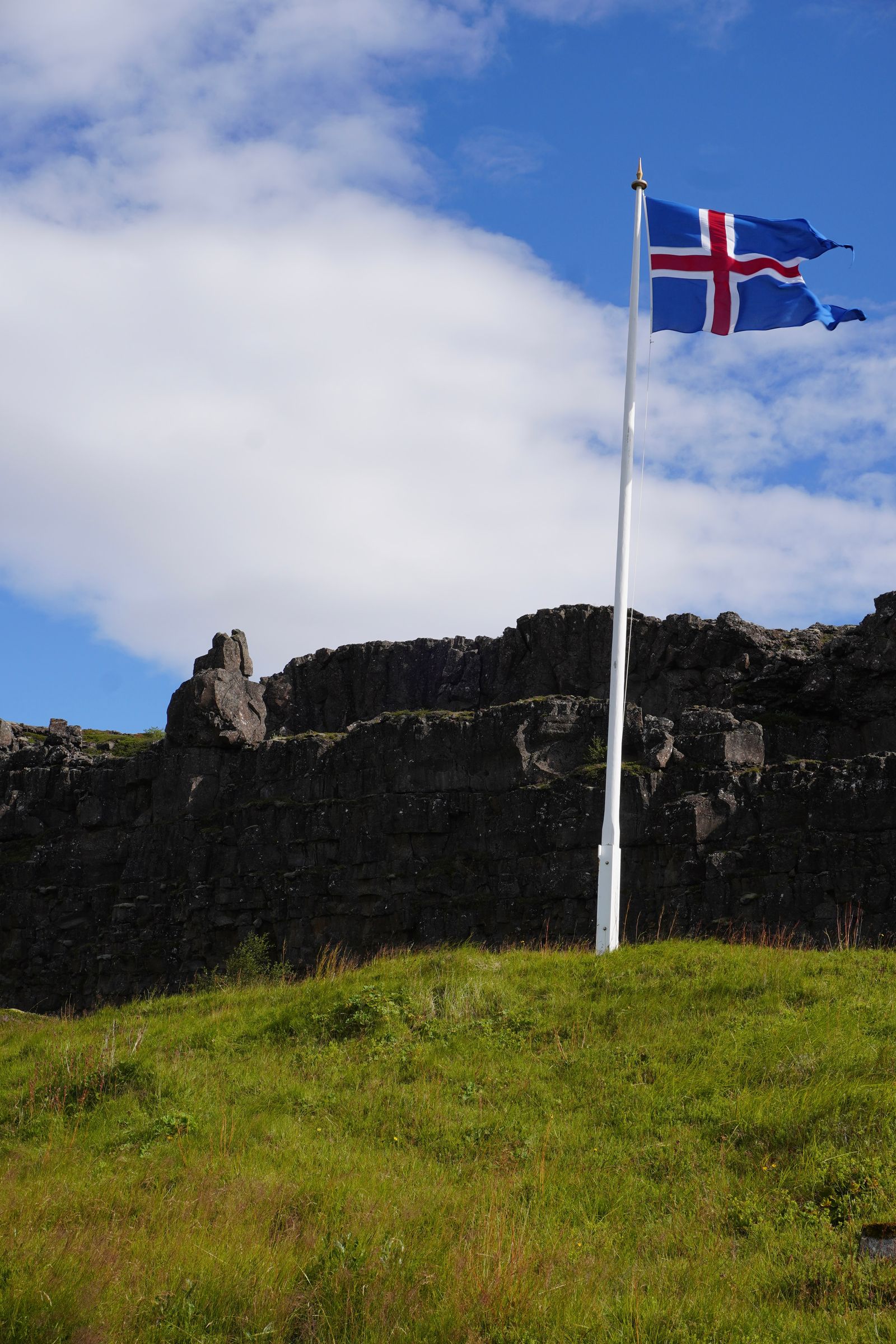 Blue flag flying above grass beside dark rock, Bláskógabyggð