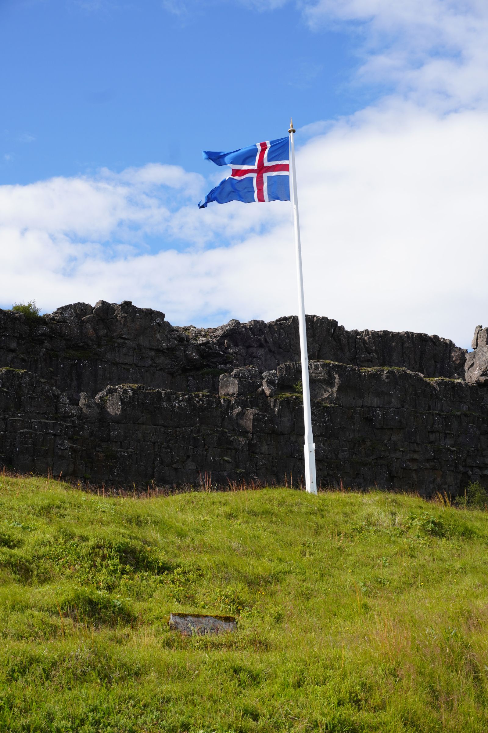 Blue flag flying above grass beside dark cliffs, Bláskógabyggð