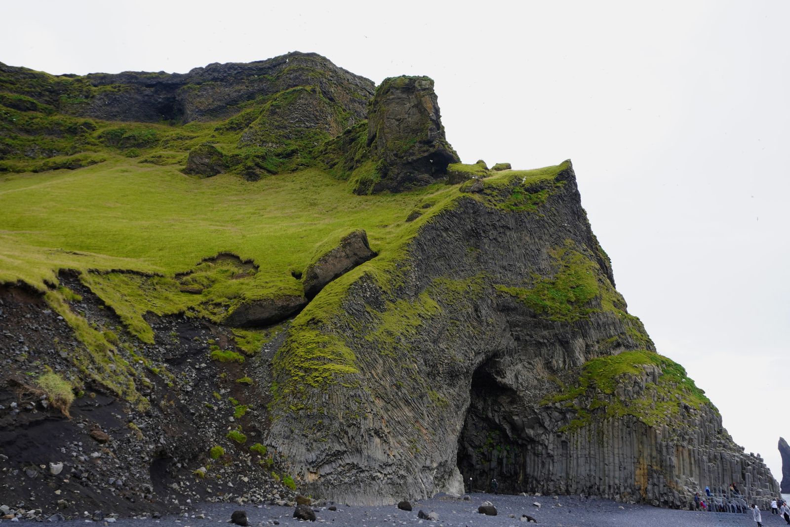 Grass-covered cliff with a cave above a dark pebble beach, Skogafoss