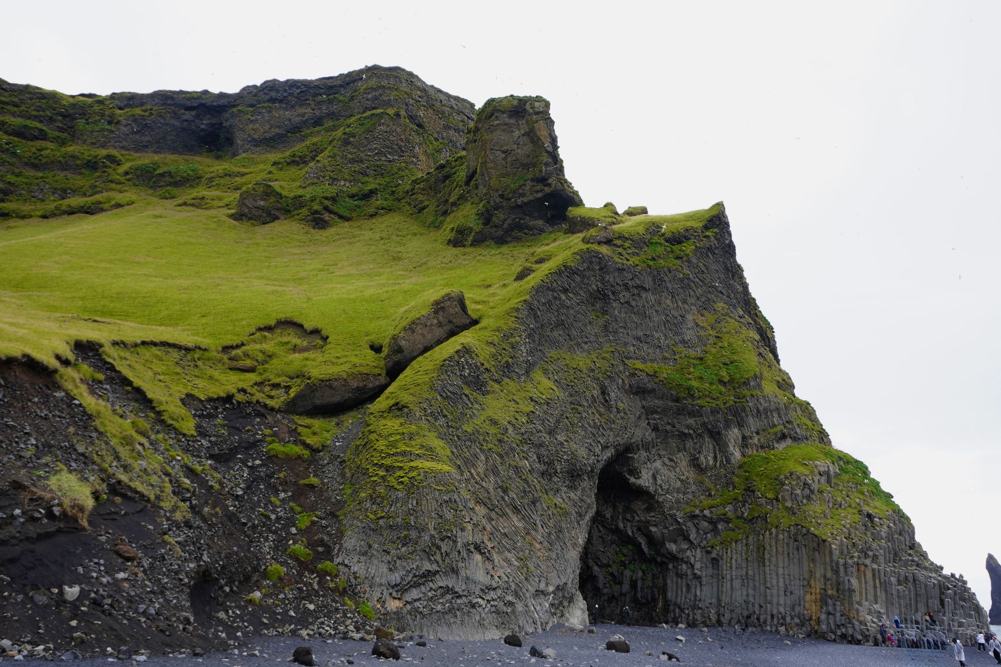 Grass-covered cliff with a cave above a dark pebble beach, Skogafoss