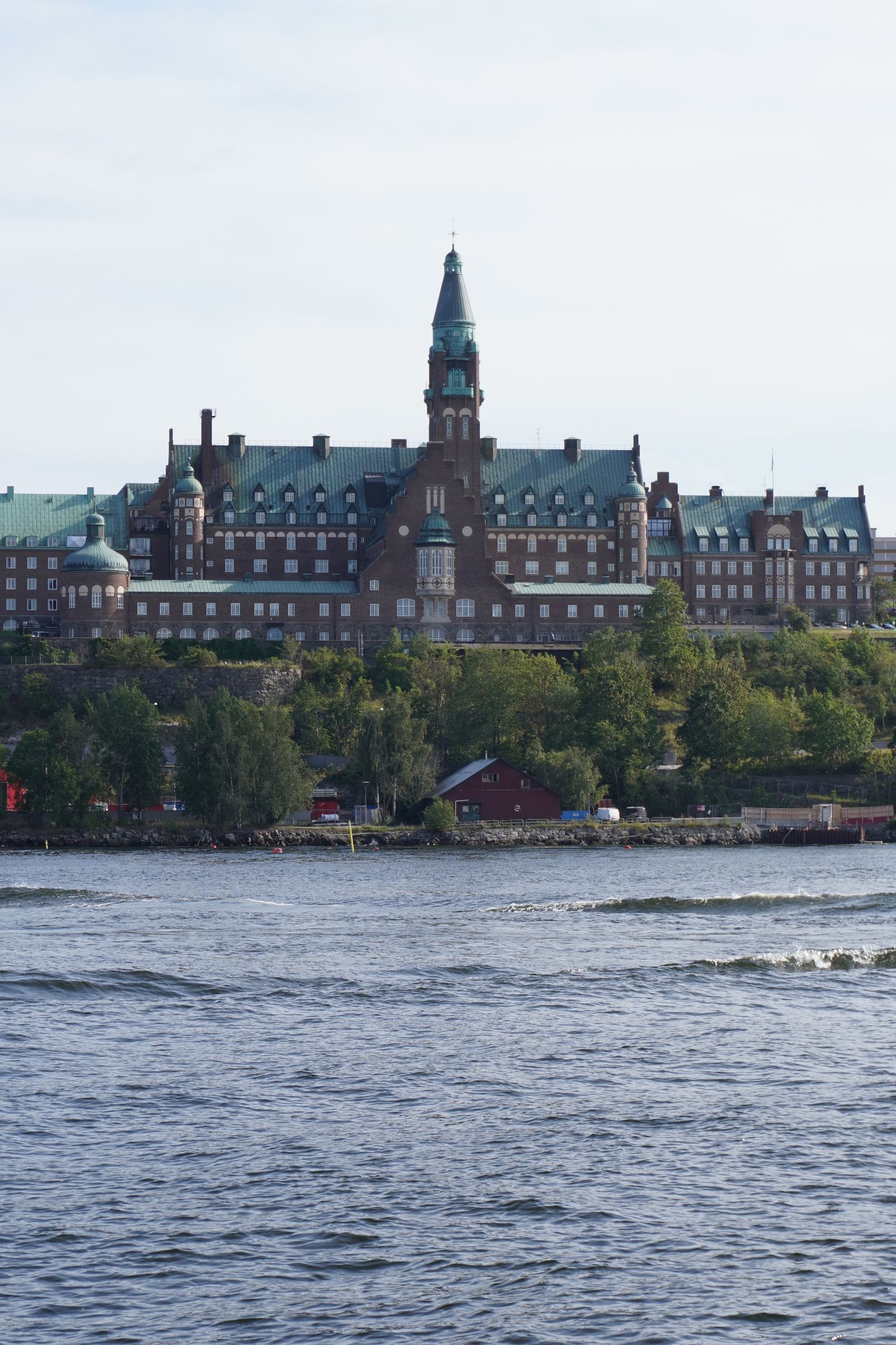 Brick building with a tall central tower above a tree-lined shore with water in front, Stockholm