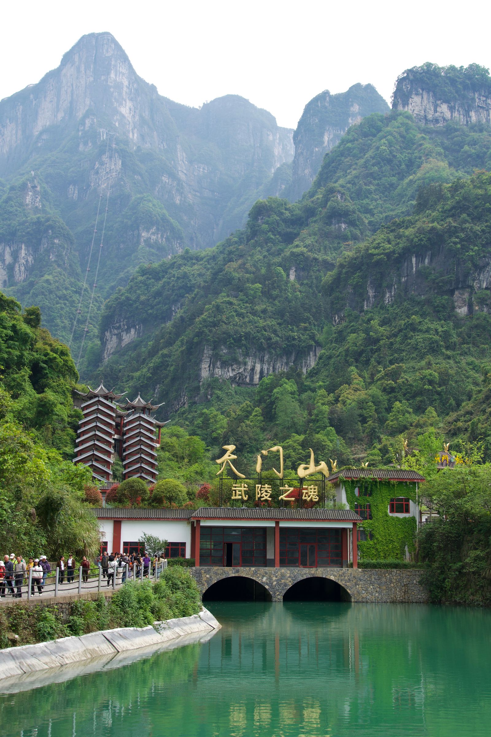 Forested mountains above buildings reflected in a green pool, Zhangjiajie