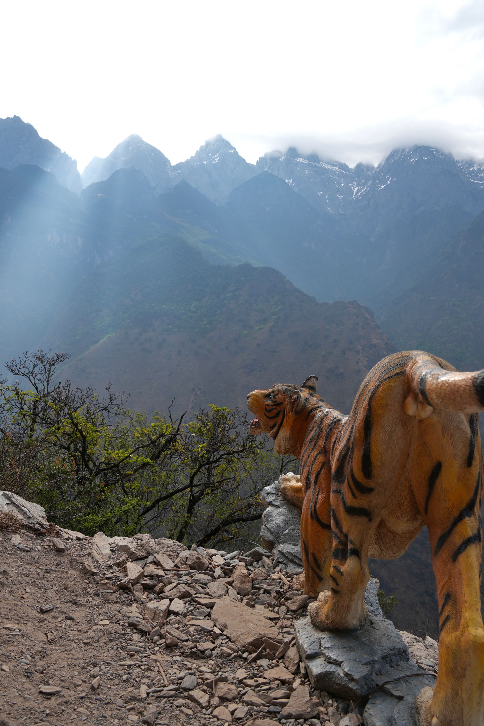Striped animal statue on rocks below snow-streaked mountains in clouds, Tiger Leaping Gorge
