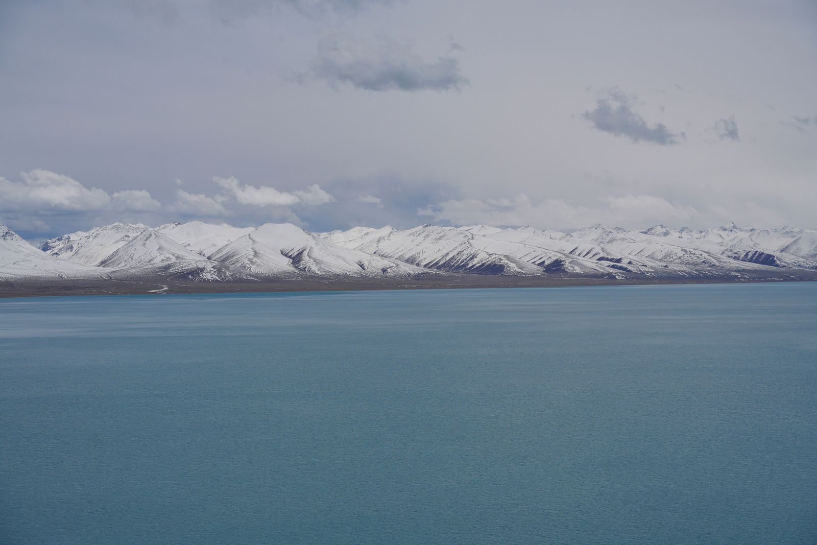 A blue lake below snow-covered mountains under gray clouds, Tibetan plateau