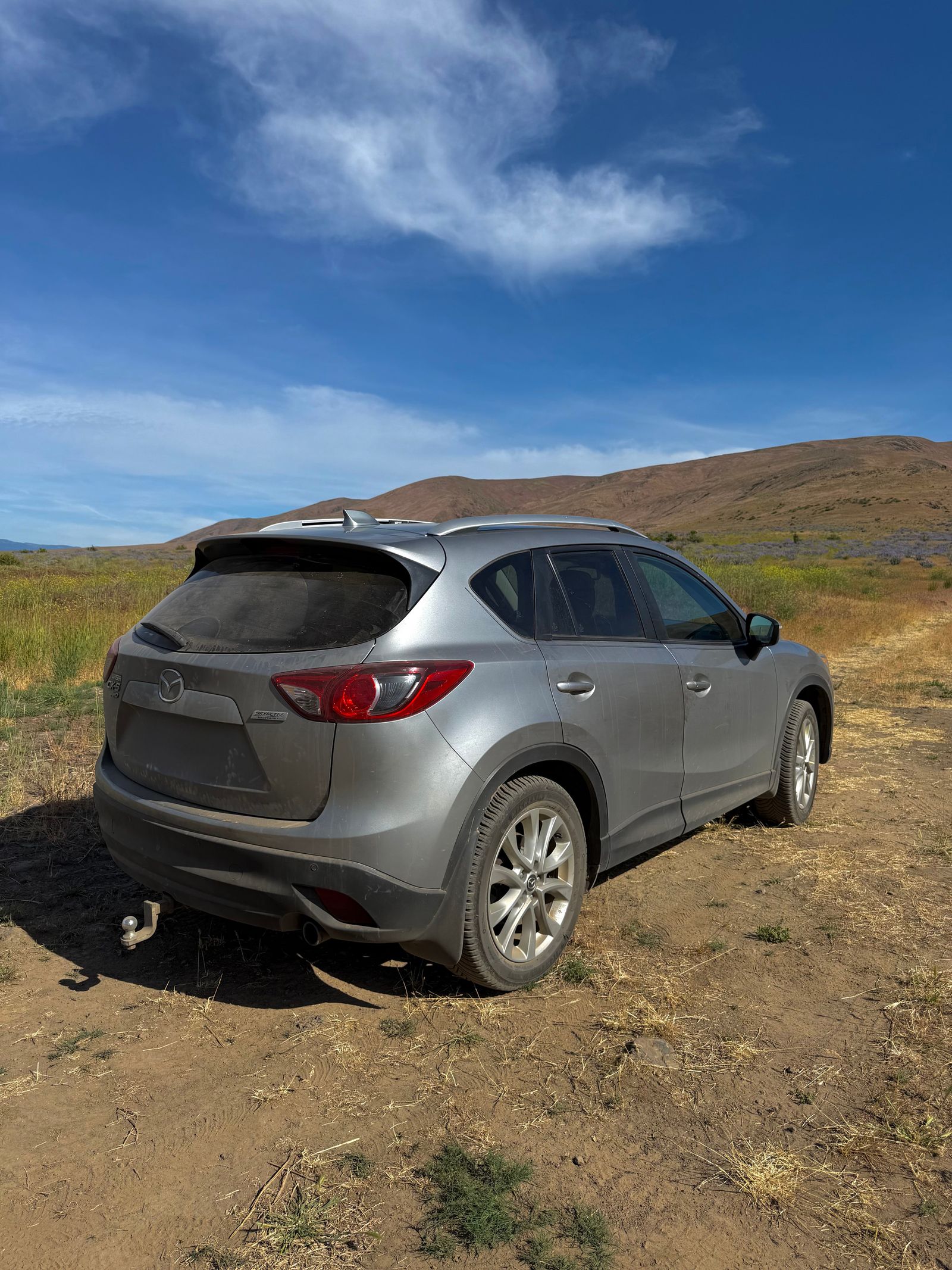 A silver SUV parked on dirt below grassy hills, shrub-steppe