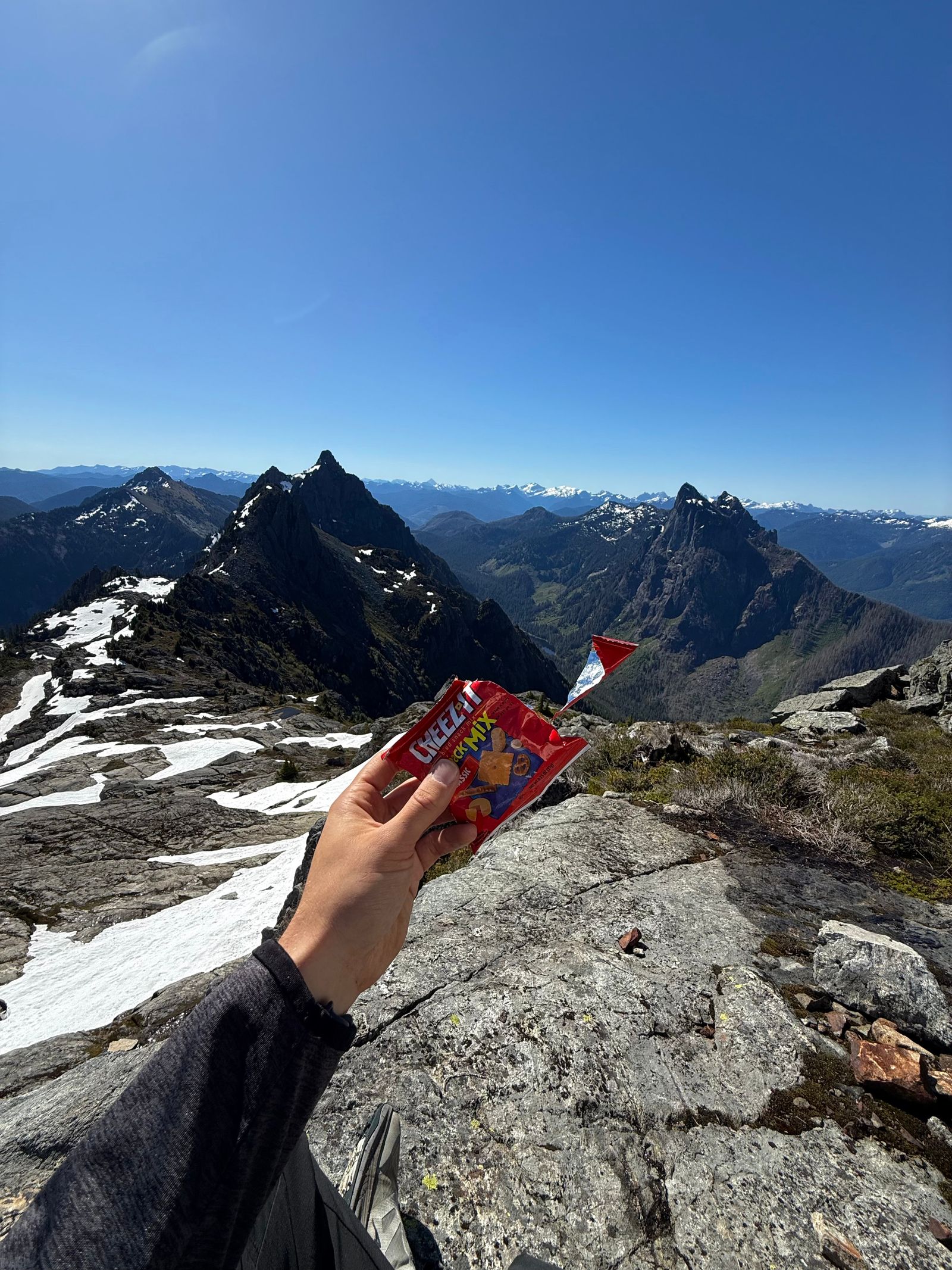 A hand holding a red snack bag below rocky peaks, Gunn Peak