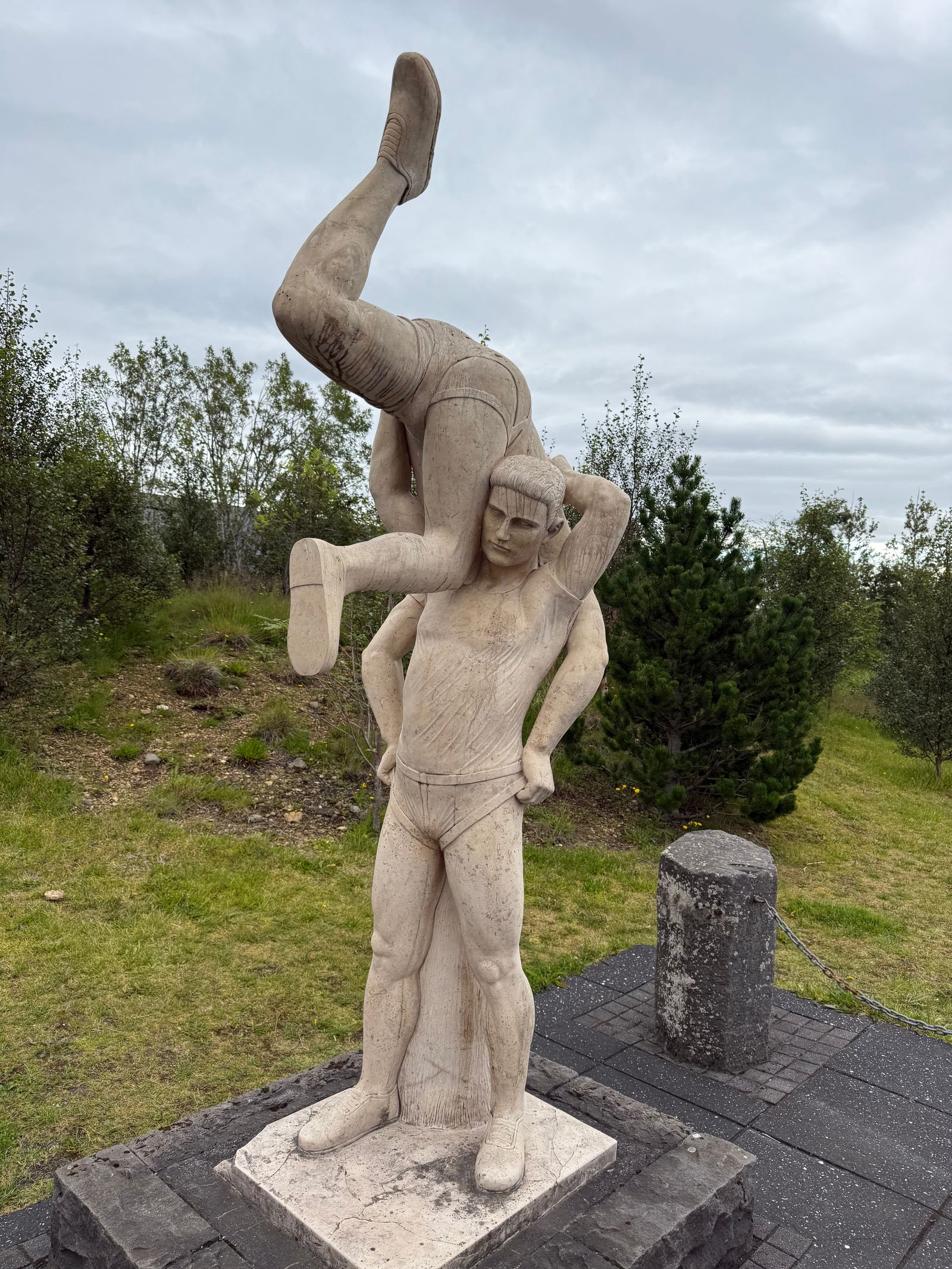 Sculpture of two figures on a stone base among trees, Bláskógabyggð