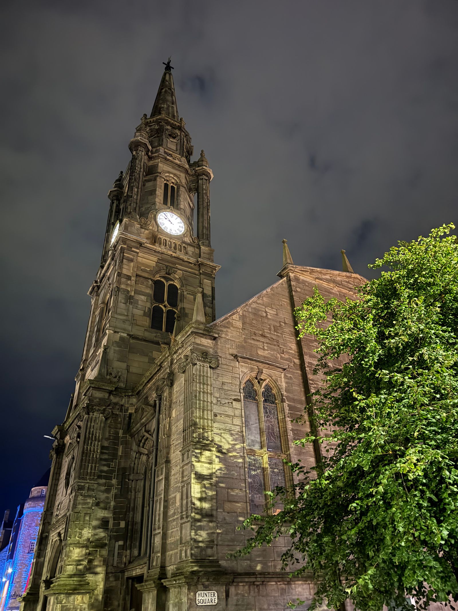 Stone church clock tower beside a leafy tree at night, Edinburgh Town